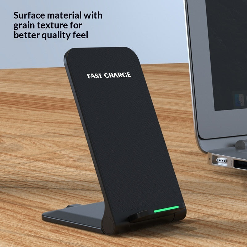 A black wireless charging pad standing upright on a wooden desk, with a laptop partially visible on the right and text in the top left reading 'Surface material with grain texture for better quality feel'. The charging pad has 'FAST CHARGE' printed on it and a small green LED indicator light at the bottom.