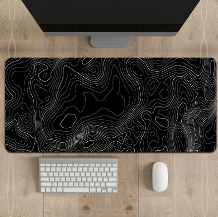 Desk setup with a large black and white topographic mouse pad, keyboard, and computer monitor on a wooden surface.