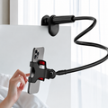 A black flexible phone mount clip with a smartphone attached, shown mounted on a white bedside table near a window with a hand reaching toward the phone holder.