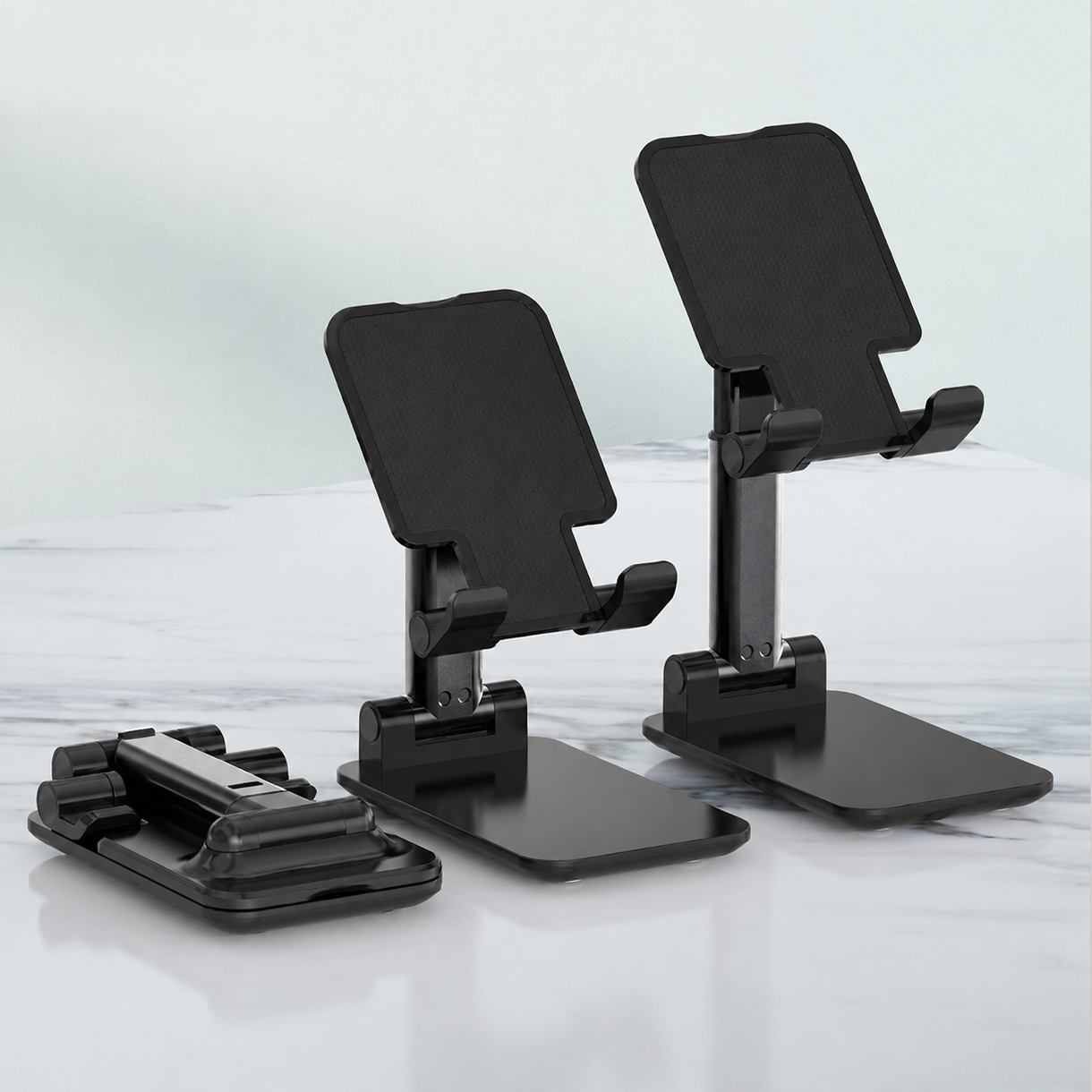 Black adjustable smartphone stands with a stand folded flat beside them, shown on a light gray marble-like surface against a pale gray background.