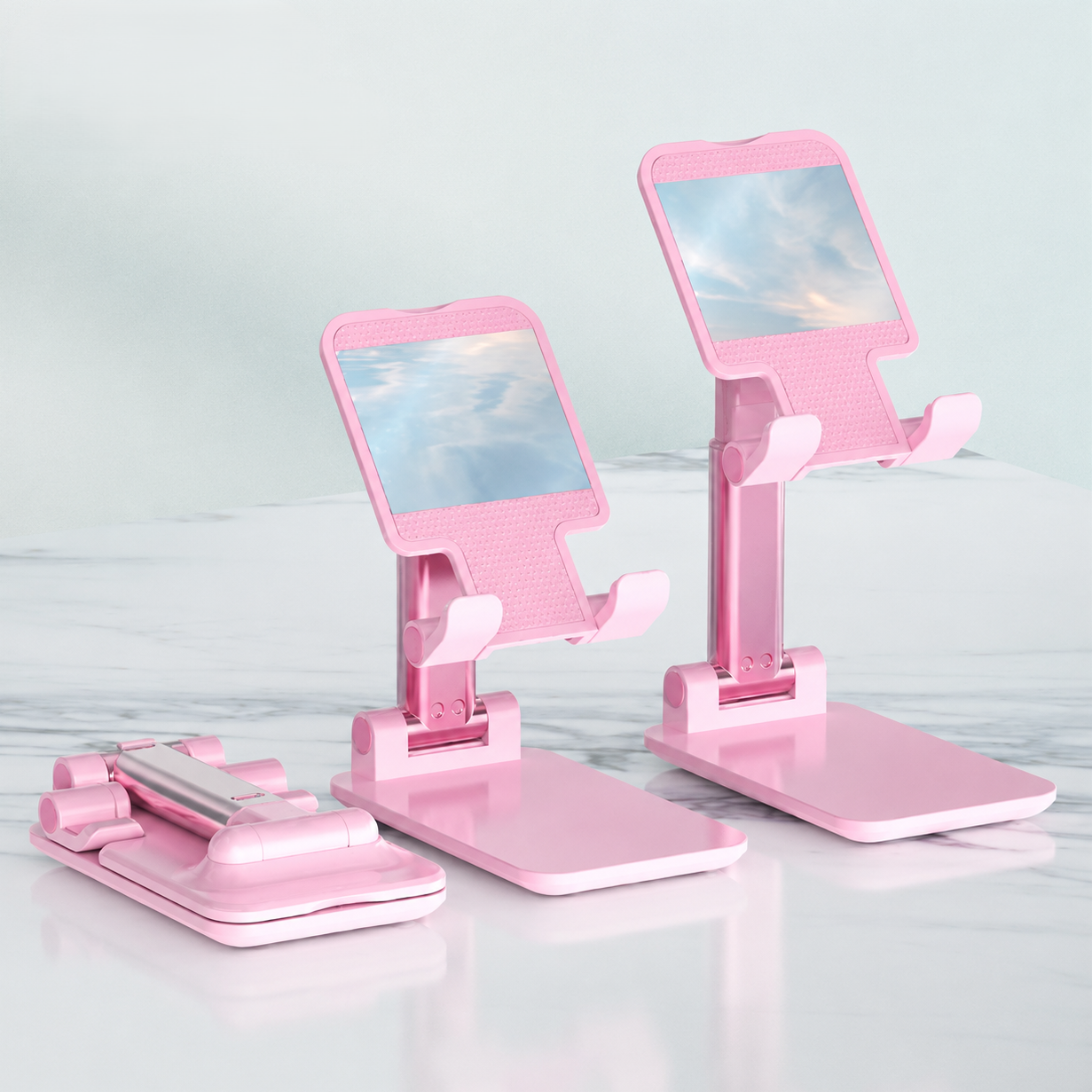 Pink foldable smartphone stands arranged on a light marble-like surface against a pale mint background. One stand is open and angled, one is open and upright, and one is closed showing the hinge mechanism.