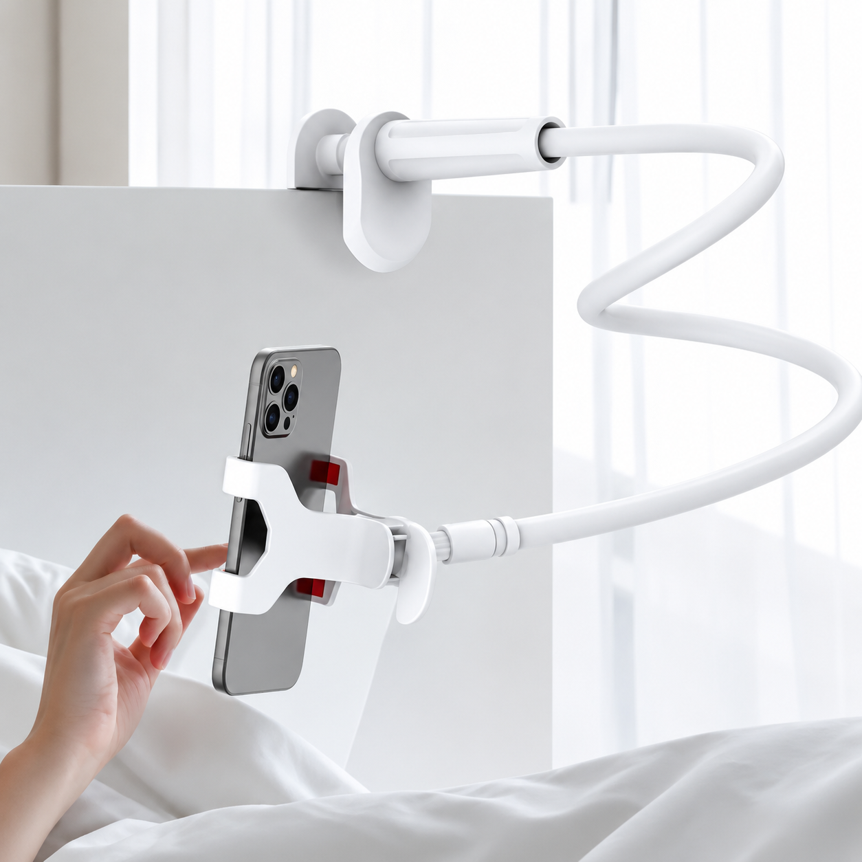 A white flexible arm phone holder mounted on a white bedside table or bed panel, holding a smartphone. A person's hand and part of a bed with white bedding are visible in a bright bedroom setting.