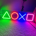 A rectangular acrylic LED neon sign featuring the PlayStation-style triangle, circle, cross, and square symbols glowing in green, red, blue, and pink, placed on a carpeted floor with colored ambient lighting.