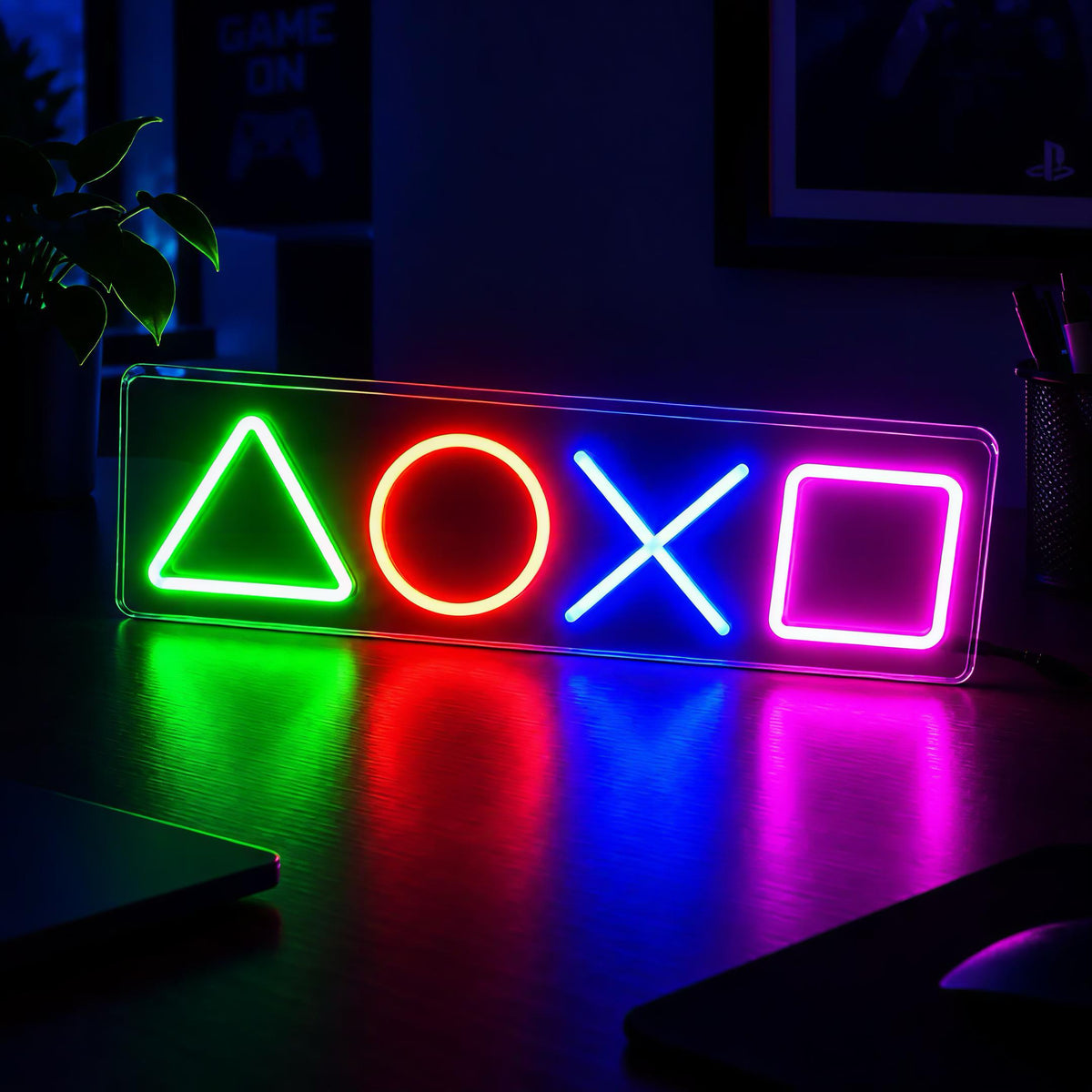 A rectangular acrylic neon sign featuring the PlayStation-style triangle, circle, cross, and square symbols glowing in green, red, blue, and pink, placed on a dark desk with colorful reflections.