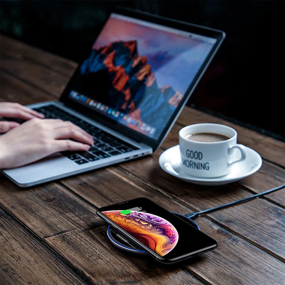 Person using a laptop with a smartphone on a wireless charger and a cup of coffee on a wooden table.