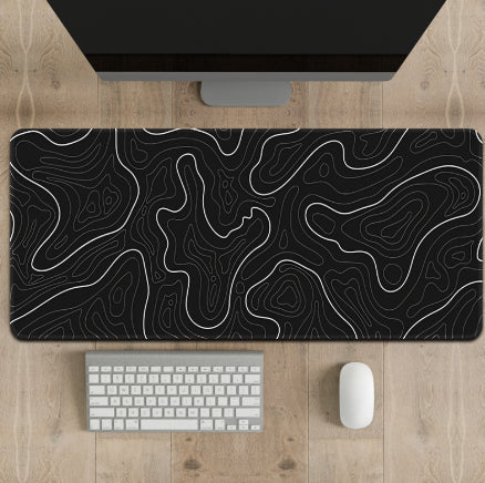 Black and white topographic desk mat on a wooden desk with a computer monitor, keyboard, and mouse.